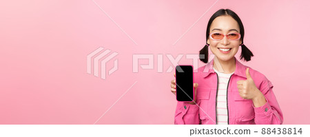 Happy stylish girl recommends application on mobile phone. Smiling asian female model showing smartphone screen and thumb up, standing over pink background 88438814