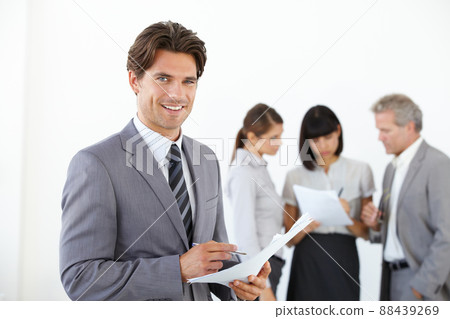 A young handsome executive holding documents while his team stands in the background. A young handsome executive holding documents while his team stands in the background. A young handsome executive holding documents while his team stands in the background. A young handsome executive holding documents while his team stands in the background. 88439269