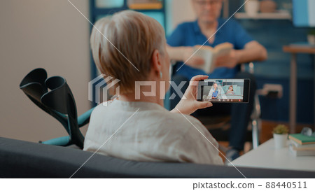 Old woman with crutches using video call on mobile phone. Senior adult talking to medic and child on online teleconference to start remote conversation. Grandmother with smartphone. 88440511