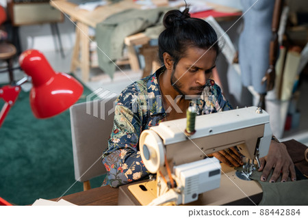 High angle shot of young fclothes designer of Indian ethnicity sitting at table using sewing machine to make dress for new collection 88442884