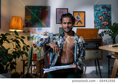 Medium portrait of young fashion designer wearing stylish casual clothes sitting on chair in living room holding pencil and papers looking at camera 88442898