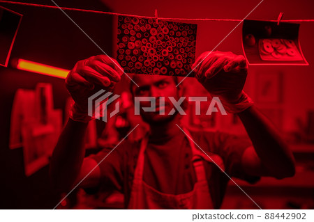 Selective focus shot of young Indian man wearing protective gloves and apron hanging wet photos on thread in darkroom 88442902