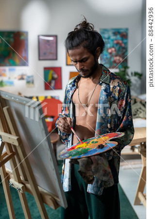 Portrait of handsome young Indian artist wearing unbuttoned shirt standing in front of easel with canvas mixing oil paints on wooden pallete 88442936