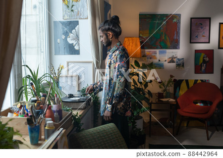 Creative young Indian man standing in living room decorated with various paintings turning on music on vinyl record player 88442964