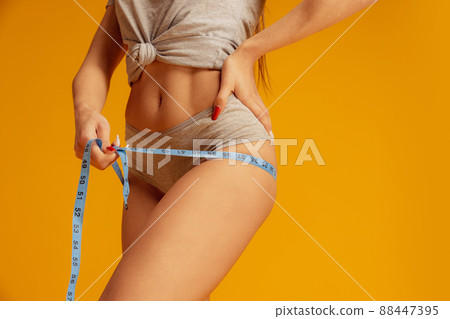 Measuring. Side view image of cropped slim tanned woman body in gray underwear on yellow studio background. 88447395