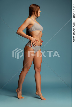 Full-length portrait of beautiful woman with perfect sportive body posing in gray cotton undewear isolated over blue studio background Full-length portrait of beautiful woman with perfect sportive body posing in gray cotton undewear isolated over blue studio background 88447445