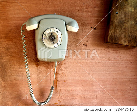 Light gray analog phone with cracked dial and dirty coiled cord hangs on pink painted stained wall 88448510