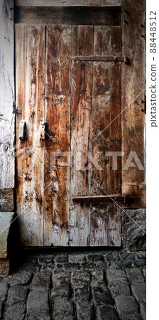 Shuttered leaning wooden door with weathered peeling brown glaze on a floor of cobblestones 88448512