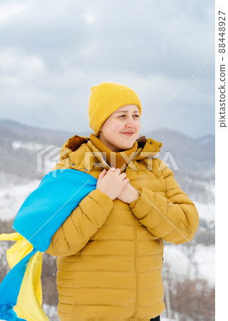 Young girl holding a blue and yellow State Flag of Ukraine against the backdrop of the Carpathian Mountains and blue sky. A woman in an orange jacket and yellow hat stands in snowy weather. 88448927