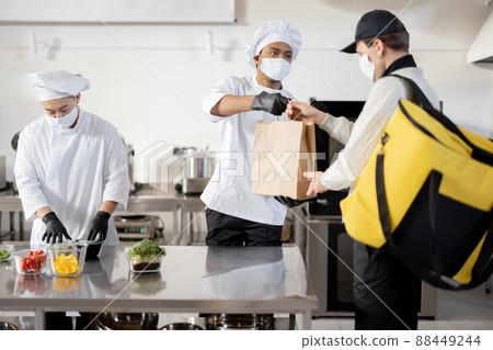 Courier waiting for an order for delivery in the kitchen with chefs preparing takeaway food Courier waiting for an order for delivery in the kitchen with chefs preparing takeaway food 88449244