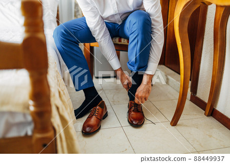 Man in a white shirt and blue trousers is tying his shoes sitting on a chair. Close-up Man in a white shirt and blue trousers is tying his shoes sitting on a chair. Close-up 88449937