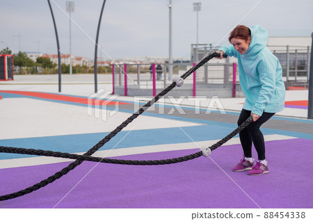 Caucasian woman in a mint sweatshirt is training with battle ropes at the sports ground.  88454338