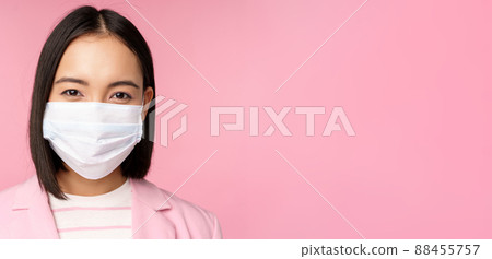 Close up portrait of japanese businesswoman in medical face mask, suit, looking at camera, standing over pink background Close up portrait of japanese businesswoman in medical face mask, suit, looking at camera, standing over pink background 88455757