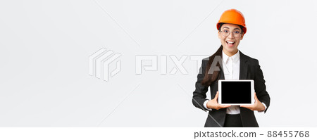 Excited and amazed smiling asian female construction engineer, architect in safety helmet and business suit, showing digital tablet screen with amazed expression, standing white background 88455768