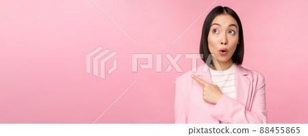 Portrait of saleswoman, korean businesswoman pointing and looking left with surprised, intrigued face expression, posing in suit over pink background 88455865