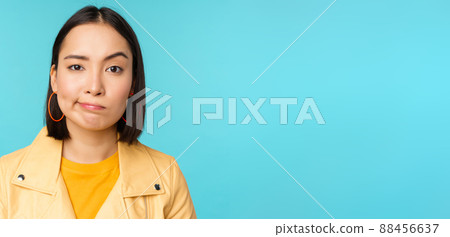 Close up of skeptical asian girl looking disappointed, staring with doubt or disbelief, grimacing at camera, standing over blue background 88456637