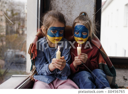 Two little girls with Ukrainian flags painted on their faces are praying for Ukraine, holding lighted candles in their hands, a gesture of faith and hope. Children against war. Two little girls with Ukrainian flags painted on their faces are praying for Ukraine, holding lighted candles in their hands, a gesture of faith and hope. Children against war. 88456765