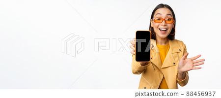 Enthusiastic asian female model, showing smartphone app interface, online store or website on mobile phone screen, standing over white background 88456937