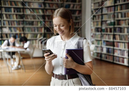 Young attractive student girl standing in library with smartphone Young attractive student girl standing in library with smartphone 88457103