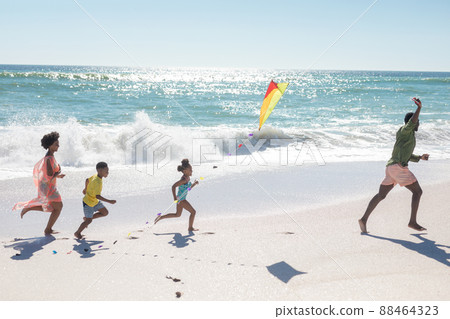 African american father running with kite ahead of family on shore at beach African american father running with kite ahead of family on shore at beach 88464323