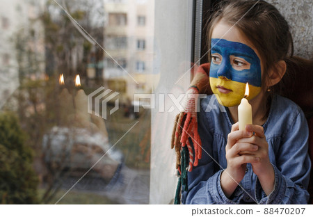 a little girl with a Ukrainian flag painted on her face praying holding a candle in her hand for the Ukrainian city of Bucha is a gesture of faith and hope. Children against war a little girl with a Ukrainian flag painted on her face praying holding a candle in her hand for the Ukrainian city of Bucha is a gesture of faith and hope. Children against war 88470207