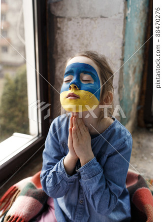 a little Ukrainian patriot girl with the flag of Ukraine on her face prays sitting at the window. Children are against the war in Ukraine 88470276