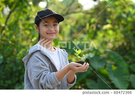Cute girl holding young plant while standing against green nature background. Saving the world, earth day concept. 88472366