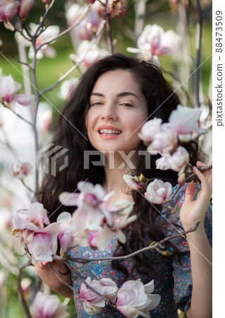 Beautiful girl with flowers of magnolia. Portrait of young smiling brunette woman under blossom magnolia tree. 88473509