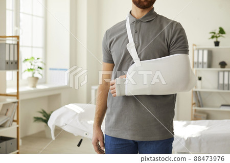 Unknown man wearing arm sling bandage on broken arm is standing in medical office. 88473976