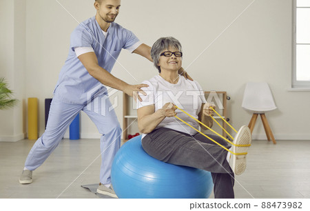 Doctor helping senior woman with osteoporosis do exercises with rubber band and fitball 88473982