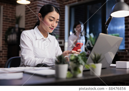 Businesswoman at work sending messages on social media platform using mobile phone. Happy smiling asian office worker sitting in office workspace while texting somebody on modern smartphone device. 88476856