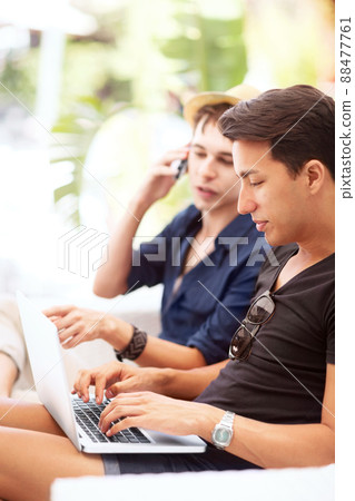 Im checking out the plcs as we speak. A young man talking on his phone while looking at his friends laptop next to him. Im checking out the plcs as we speak. A young man talking on his phone while looking at his friends laptop next to him. 88477761