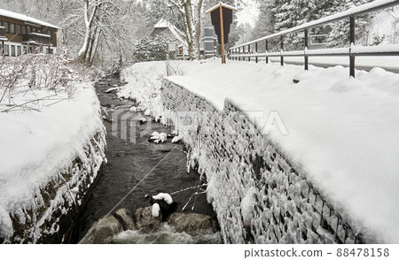 Brook with streambed forced into walls flows through snowy village in winter in Harz mountains, Germany Brook with streambed forced into walls flows through snowy village in winter in Harz mountains, Germany 88478158