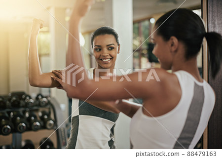 Looking good. A young woman flexing her muscles in front of the mirror at the gym. Looking good. A young woman flexing her muscles in front of the mirror at the gym. 88479163
