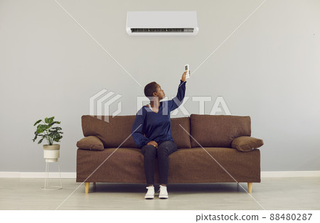 Young african woman switching on air conditioner with remote control at home. AC system concept. 88480287