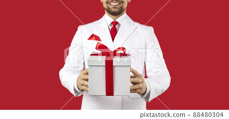 Young man in a suit standing in the studio and holding a white gift box tied with a red bow 88480304