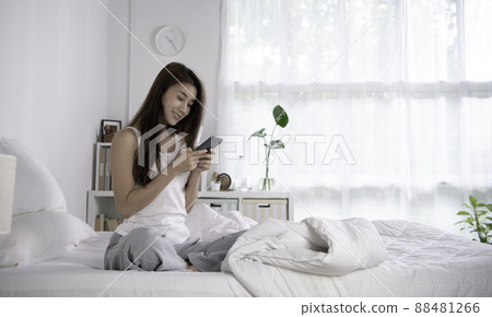 Smiling young woman relax in bedroom wake up in the morning texting messaging on phone. 88481266