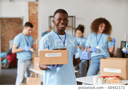 Portrait of african american young male volunteer in blue uniform, protective gloves smiling, showing thumbs up and holding cardboard box for Donation Portrait of african american young male volunteer in blue uniform, protective gloves smiling, showing thumbs up and holding cardboard box for Donation 88482172