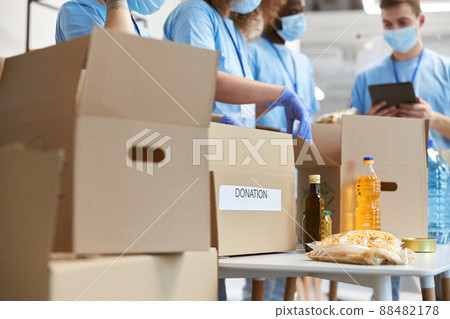 Close up of donation box. Volunteers in protective masks and gloves sorting and packing food in cardboard boxes, working on donation project indoors Close up of donation box. Volunteers in protective masks and gloves sorting and packing food in cardboard boxes, working on donation project indoors 88482178