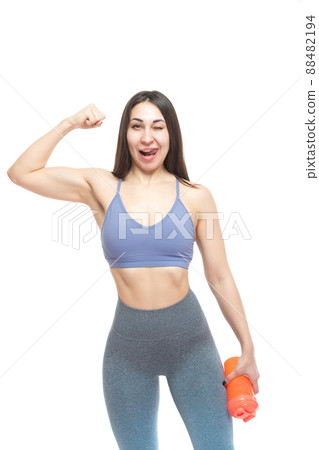 A beautiful, athletic, slender and cheerful woman demonstrates a muscular biceps arm, winks and holds a bottle of water. Lifestyle concept with sports and gym. Isolated on white background. A beautiful, athletic, slender and cheerful woman demonstrates a muscular biceps arm, winks and holds a bottle of water. Lifestyle concept with sports and gym. Isolated on white background. 88482194
