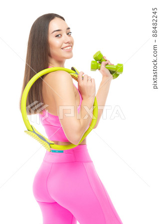 A beautiful, athletic, slim, smiling and cheerful woman in a pink tracksuit is standing with a yellow pilates hoop and green dumbbells. Lifestyle concept with sports and gym. Isolated on white 88482245
