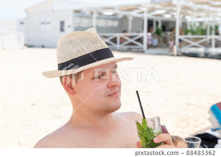 Portrait of young adult caucasian man wearing hat enjoy having fun drinking fresh mojito cocktail and sunbathing on sand beach on hot summer day. Seaside or ocean leisure vacation concept 88483362