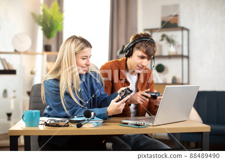 Young happy couple playing video games at home, sitting at the table with laptop and using joysticks 88489490