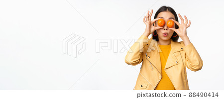 Funny asian girl holding tangerine on eyes and smiling, making playful grimaces, standing over white background Funny asian girl holding tangerine on eyes and smiling, making playful grimaces, standing over white background 88490414
