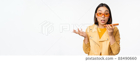 Image of happy asian girl talking on speakerphone, recording, translating her voice with mobile phone app, talking in smartphone dynamic, standing over white background 88490660