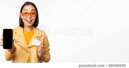 Online shopping and people concept. Stylish asian woman showing mobile phone screen and credit card, smartphone application, standing over white background Online shopping and people concept. Stylish asian woman showing mobile phone screen and credit card, smartphone application, standing over white background 88490695