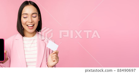 Smiling korean businesswoman in suit, showing mobile phone screen, credit card, showing online banking application interface, pink background Smiling korean businesswoman in suit, showing mobile phone screen, credit card, showing online banking application interface, pink background 88490697
