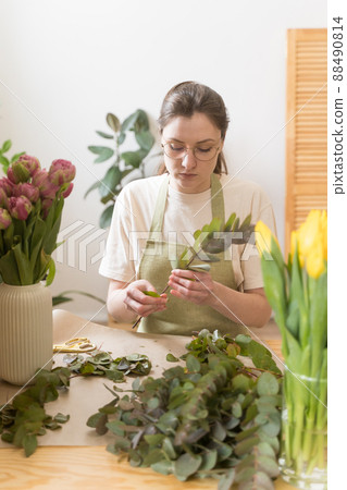 Confident florist businesswoman works with flowers. A woman in a green apron and glasses looks at a branch of eucalyptus Confident florist businesswoman works with flowers. A woman in a green apron and glasses looks at a branch of eucalyptus 88490814