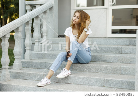 Modern attractive stylish young woman in a fashionable t-shirt in blue jeans in white sneakers resting sitting on the steps of a vintage building. Cute European girl enjoying summer day outdoors. Modern attractive stylish young woman in a fashionable t-shirt in blue jeans in white sneakers resting sitting on the steps of a vintage building. Cute European girl enjoying summer day outdoors. 88490883