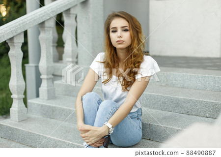 Beautiful young pretty woman with a stylish hairstyle in a trendy white t-shirt in stylish blue jeans is sitting on the vintage steps of the hotel outdoors. Modern european girl enjoying summer day. 88490887